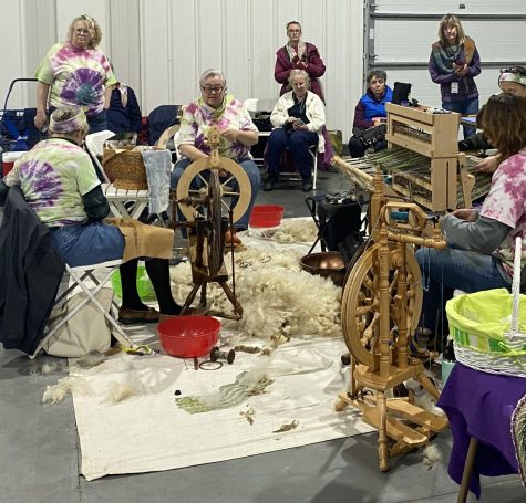 Group of women sitting in a circle. 2 sitting in front of a wheel, spinning. 1 in front of a loom.